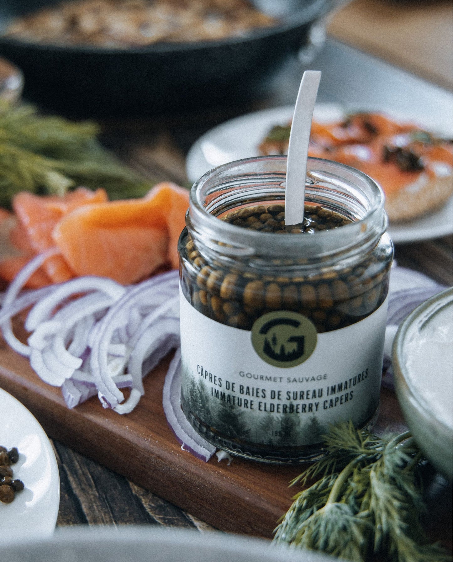 open jar of elderberry capers seen in situ on a wooden board alongside sliced red onion, smoked salmon, and dill