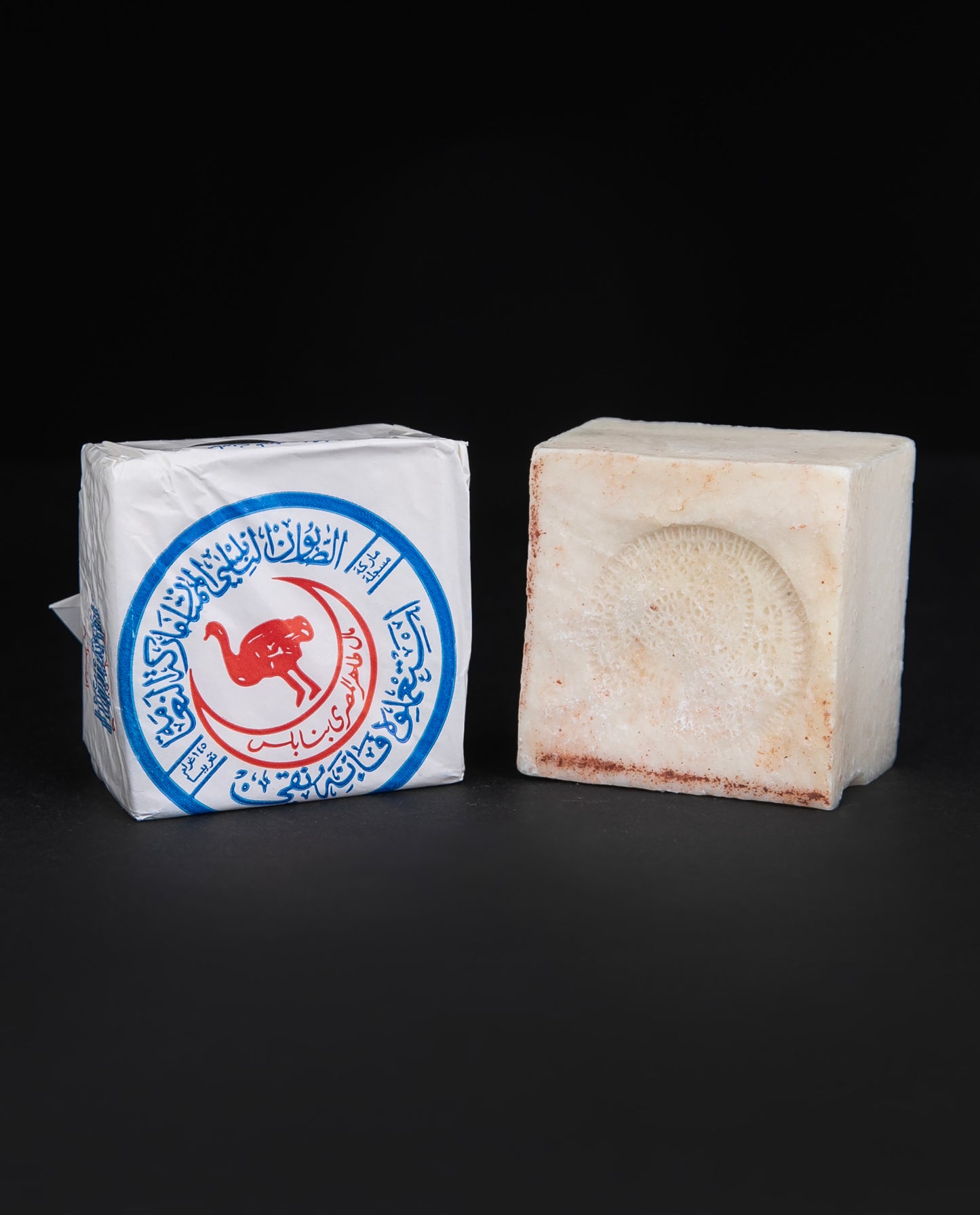 Wax paper-wrapped olive oil soap, sitting next to an unwrapped bar of soap on black background. The bar of soap is stamped with an ostrich seal.