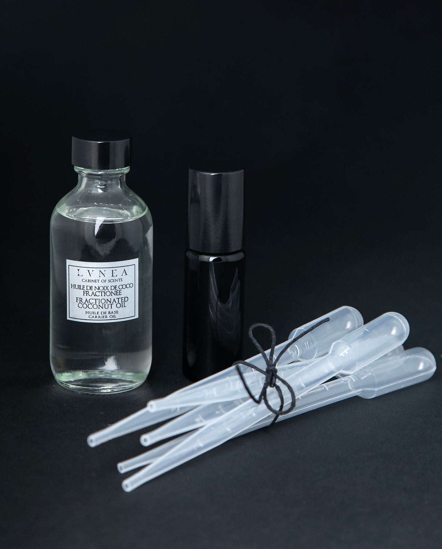 Clear 60ml glass bottle of fractionnated coconut oil, a 10ml black glass roller, and 5 clear plastic pipettes bundled together with a piece of black twine.