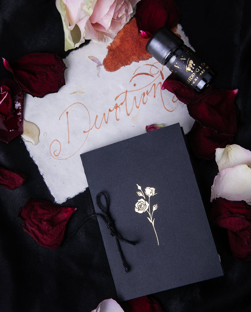  LVNEA's "Bleeding Heart" stationery set including a hand-bound scented booklet and scented ink in black glass vessel with gold foil. The set is seen from above and surrounded by scattered rose petals, and a piece of natural paper with the word "Devotion" written in red ink.