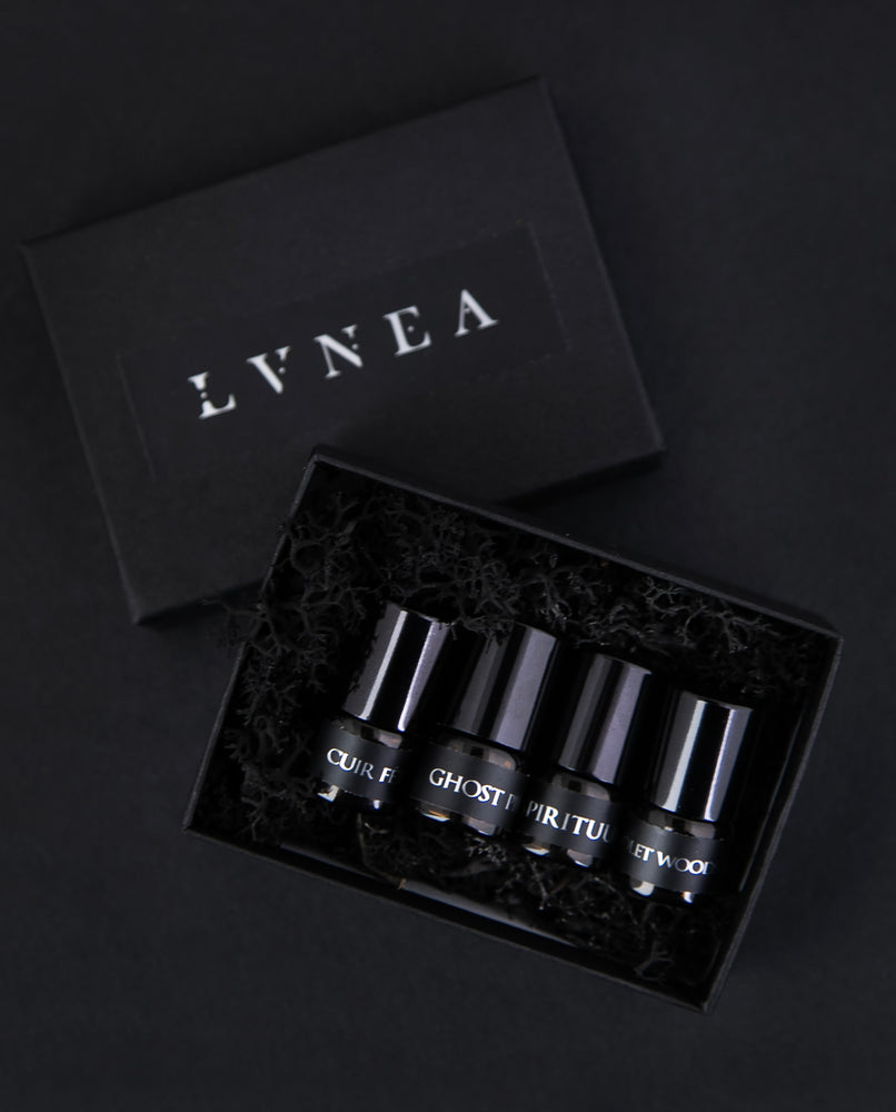 Set of 4 1.25 ml glass sample vials of LVNEA oil perfume. Shown are Cuir Fétiche, Ghost Pine, Spirituum, and Violet Woods. They are in a black box filled with moss, on a black background.