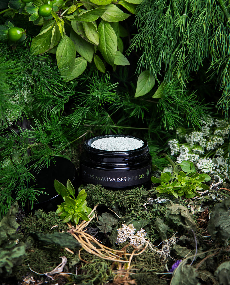 Opened black glass jar of LVNEA's limited edition Les Mauvaises Herbes powdered clay face mask, peeking out from a dense forest of herbs and vegetation.