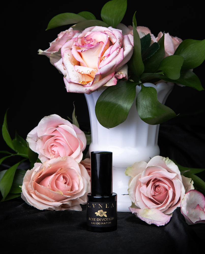 15ml black glass bottle of LVNEA's "Rose Devotion" limited edition eau de parfum, against black satin and surrounded by blush-coloured roses and greenery