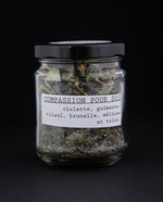Clear glass jar of Blueberryjams' "Self Compassion" herbal tea blend against black background, french label is visible and reads "Compassion pour soi"