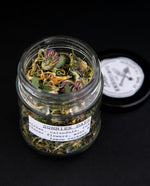 Open jar of "Sunny Days" herbal tea blend, colourful calendula, red clover, and violet flowers are visible