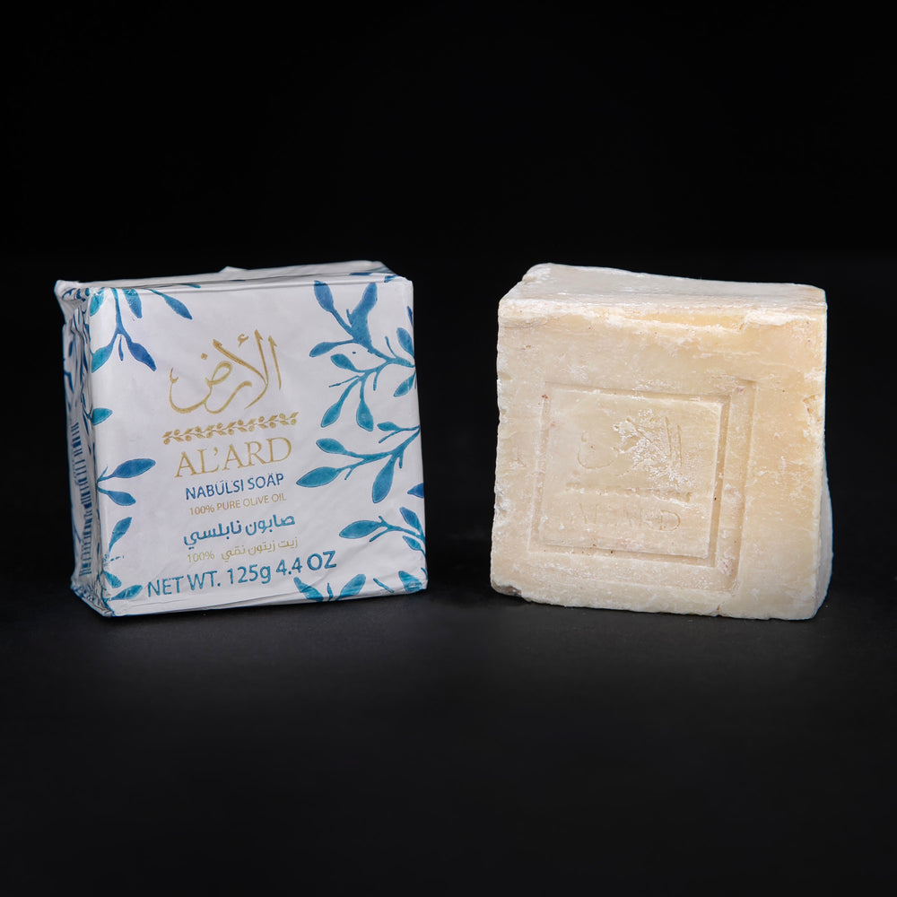 
                      
                        Wax paper-wrapped bar of olive oil soap next to unwrapped bar of soap. sitting on black background. The soap is staped with the Al'Ard logo.
                      
                    