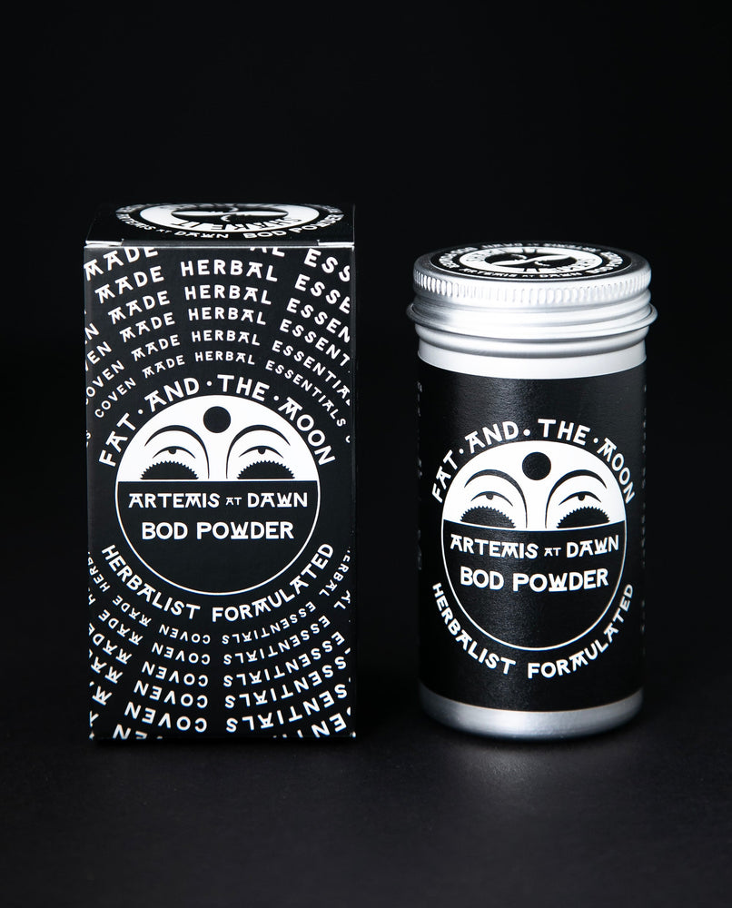 Metal tin of Fat and the Moon body powder next to its box. The label on the tin is black and white with an illustration of a half moon peeking over the logo.