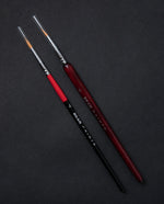 Two paintbrushes sitting side by side on a black background. One has a thin red and black handle, the other has a slightly thicker wooden handle stained red.