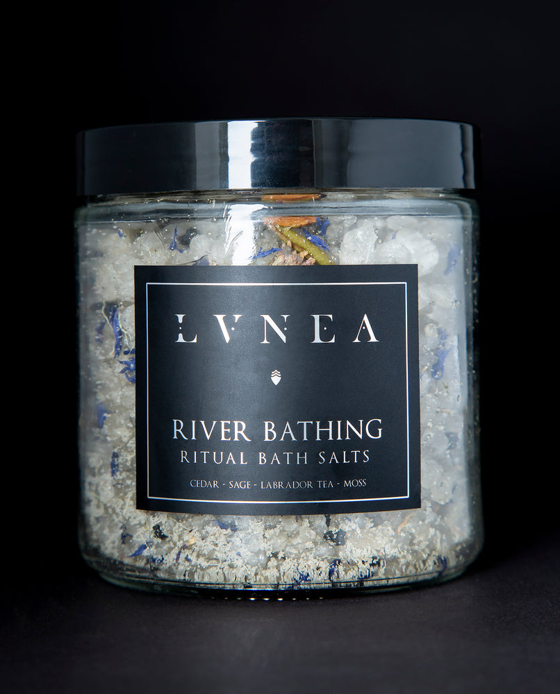 A clear 16 ounce jar filled to the brim with a cooling bath salt blend by natural / botanical perfume and beauty brand LVNEA. The label reads RIVER BATHING RITUAL BATH SALTS and highlights notes of hinoki cedar, and clary sage essential oils. The salts are a blend of small and large coarse sea salts, icelandic clay, blue cornflower petals, labrador tea buds, and strands of usnea (tree moss). 