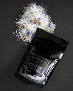 A 4 ounce  filled to the brim with a calming bath salt blend by natural / botanical perfume and beauty brand LVNEA. The label reads FOREST BATHING RITUAL BATH SALTS and highlights notes of silver fir, black spruce, pine, and vetiver essential oils. The salts are a blend of small and large coarse sea salts, french green clay, and cedar leaves.