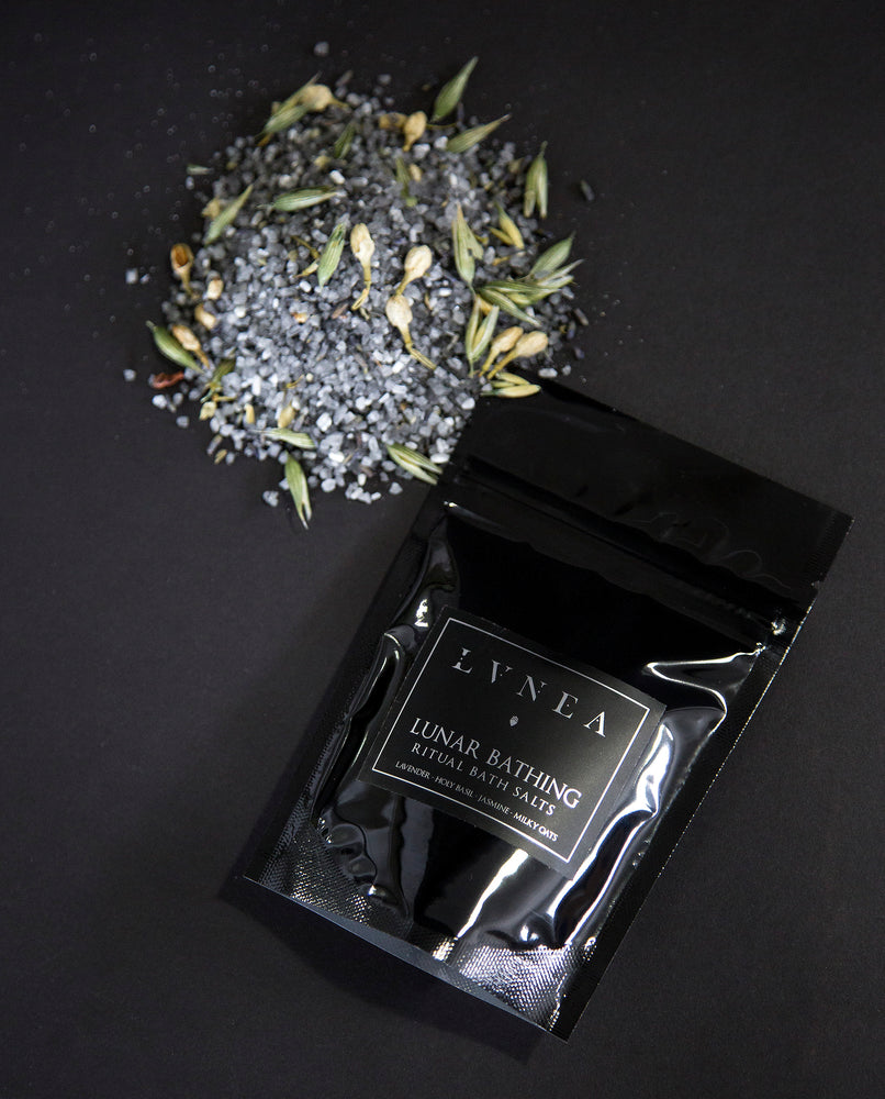 4oz black resealable pouch of LVNEA's Lunar Bathing bath salts with a mound of charcoal-hued salts speckled with milky oat tops, jasmin buds and lavender next to it