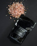 4oz black resealable pouch of LVNEA's Fire Bathing bath salts with a mound of rose-hued salt next to it