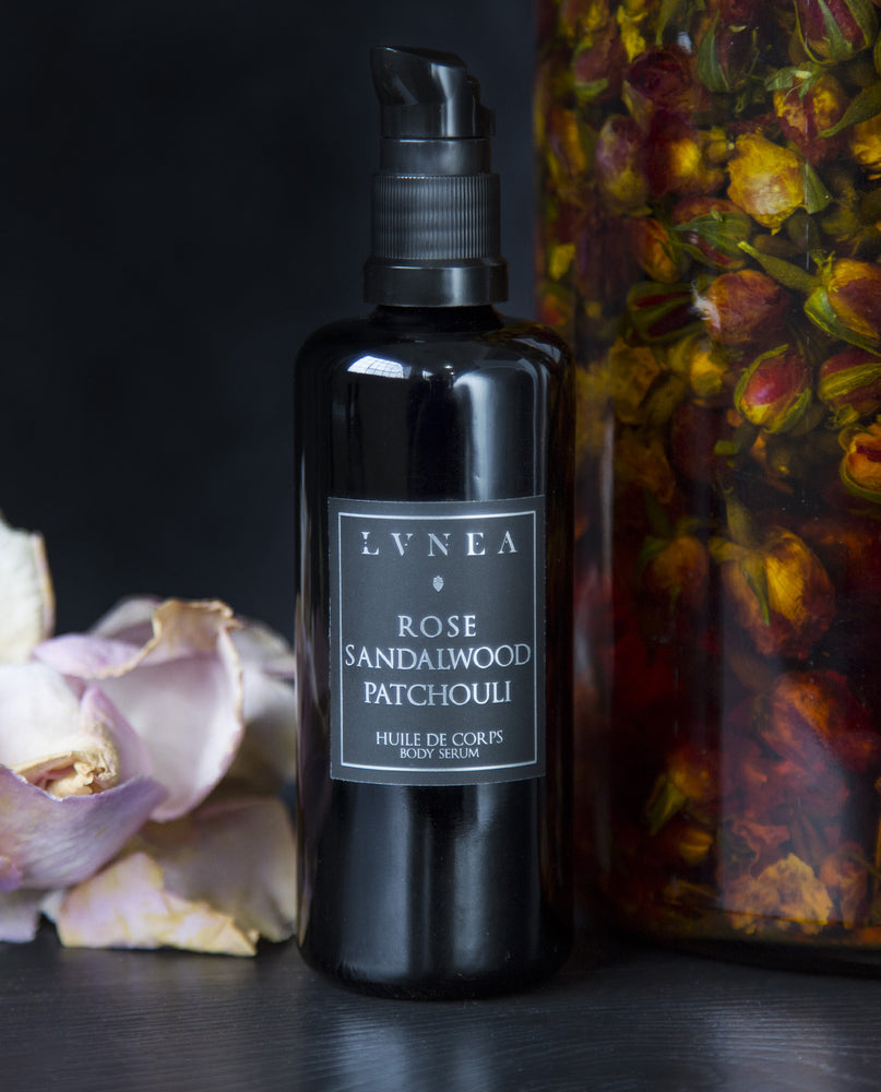 100ml black bottle of LVNEA's Rose, Sandalwood and Patchouli body serum next to a jar of rose buds infusing in oil