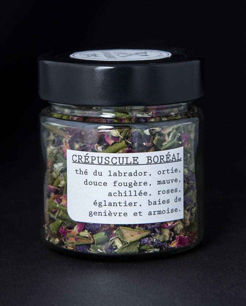 Clear glass jar with black lid containing herbal tea blend on black background. The label reads "Crépuscule Boréal"