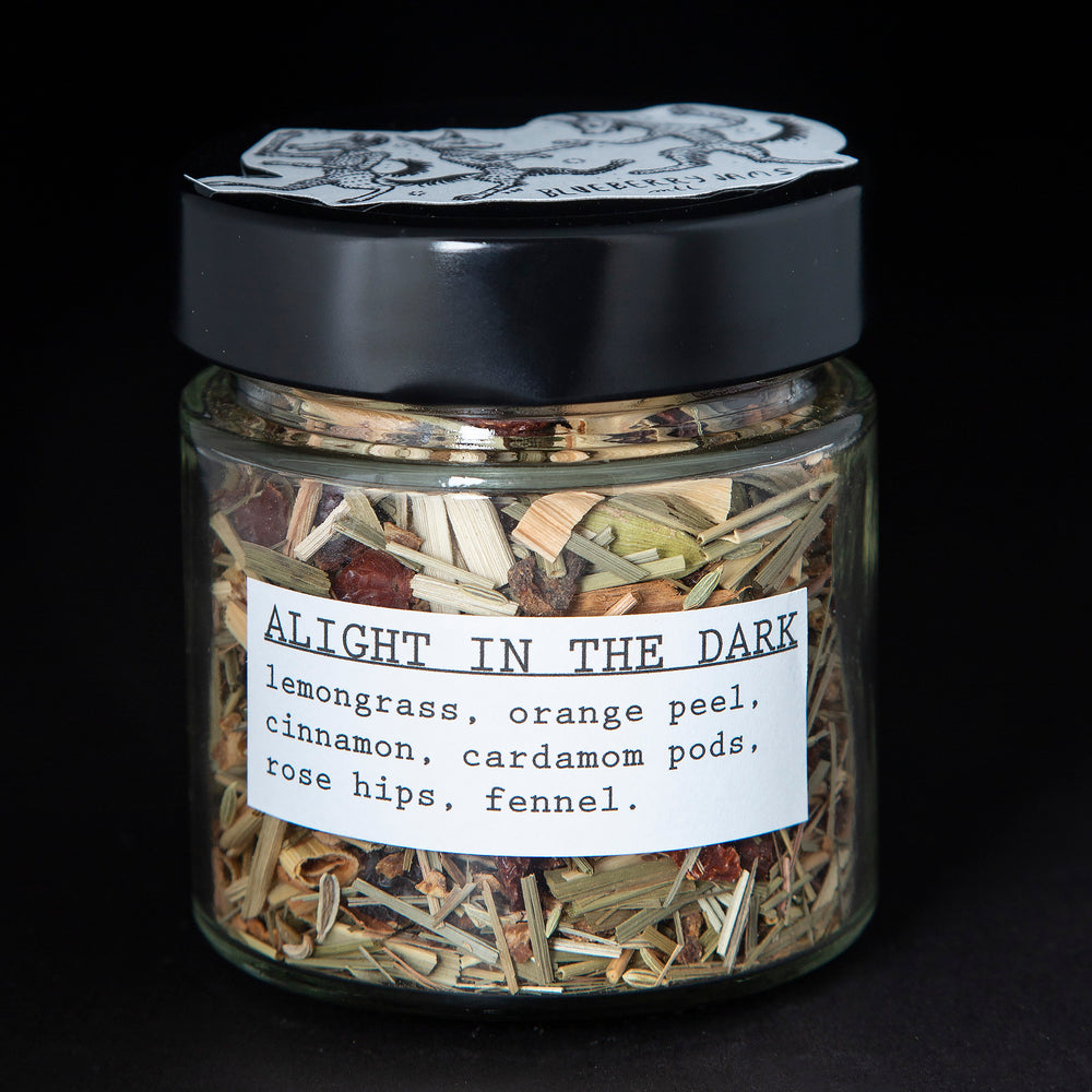 Clear glass jar with black screw on top containing blueberryjams' "Alight In The Dark" herbal tea blend. Presented on black background.