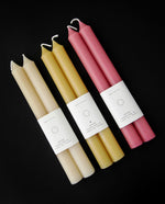 3 pairs of beeswax taper candles on black background. They are cream, yellow, and pink, respectively.
