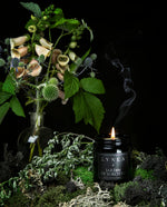 A staged photo of an herbal, aromatic artisanal and naturally scented vegan soy wax candle with a cotton wick by natural and botanical perfume and apothecary brand LVNEA with notes of mugwort, tomato leaf, moss, myrrh, and patchouli,. Hand-poured into a resealable 8 ounce / 250 ml black glass jar with violet undertones, the label reads JARDIN DE SORCIÈRE essential oil candle. The candle is lit and displayed on a bed of moss, with a curious looking bouquet of native garden flowers in the background.
