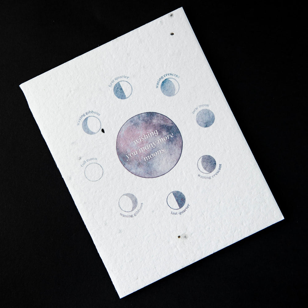 White greeting card featuring blue and purple watercolour illustrations of the moon's phases. The card reads "wishing you many more moons"