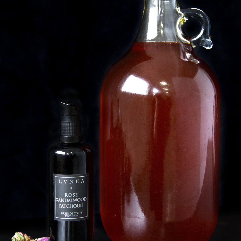 
                      
                        Black bottle of LVNEA's Rose, Sandalwood and Patchouli body serum standing next to large clear glass jug filled with amber-coloured oil
                      
                    