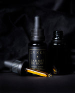 20ml black glass bottle with a black label reading "Larmes de Rose Facial Serum" in gold. There is a black unlabelled bottle next to it with its top removed, revealing a dropper filled with golden liquid.